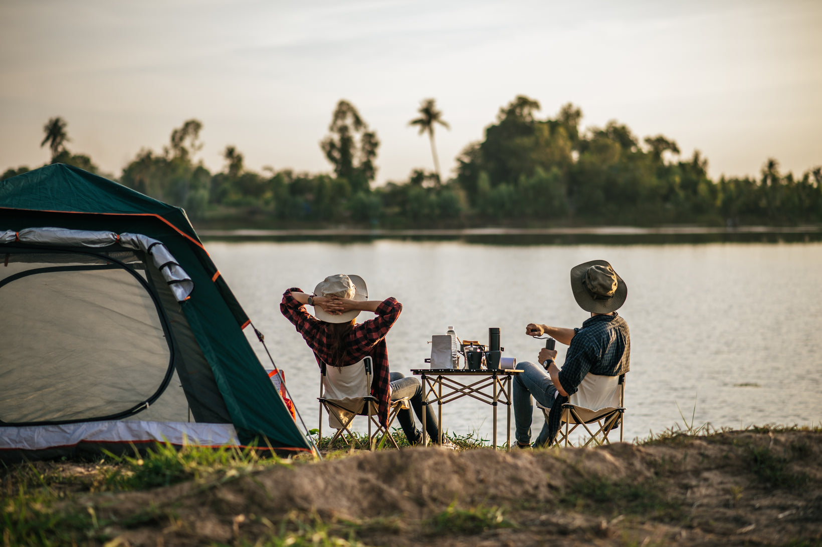 Two people camping by a lake, sitting at a table with a tent nearby.