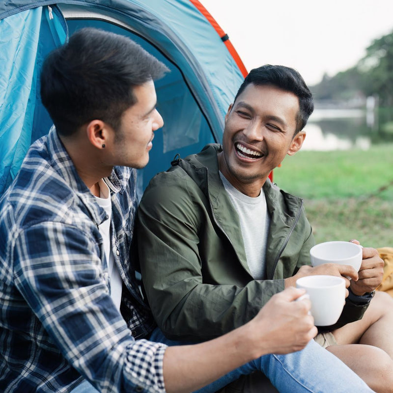 Two men sitting in front of a blue tent, enjoying a cup of coffee in a natural setting.